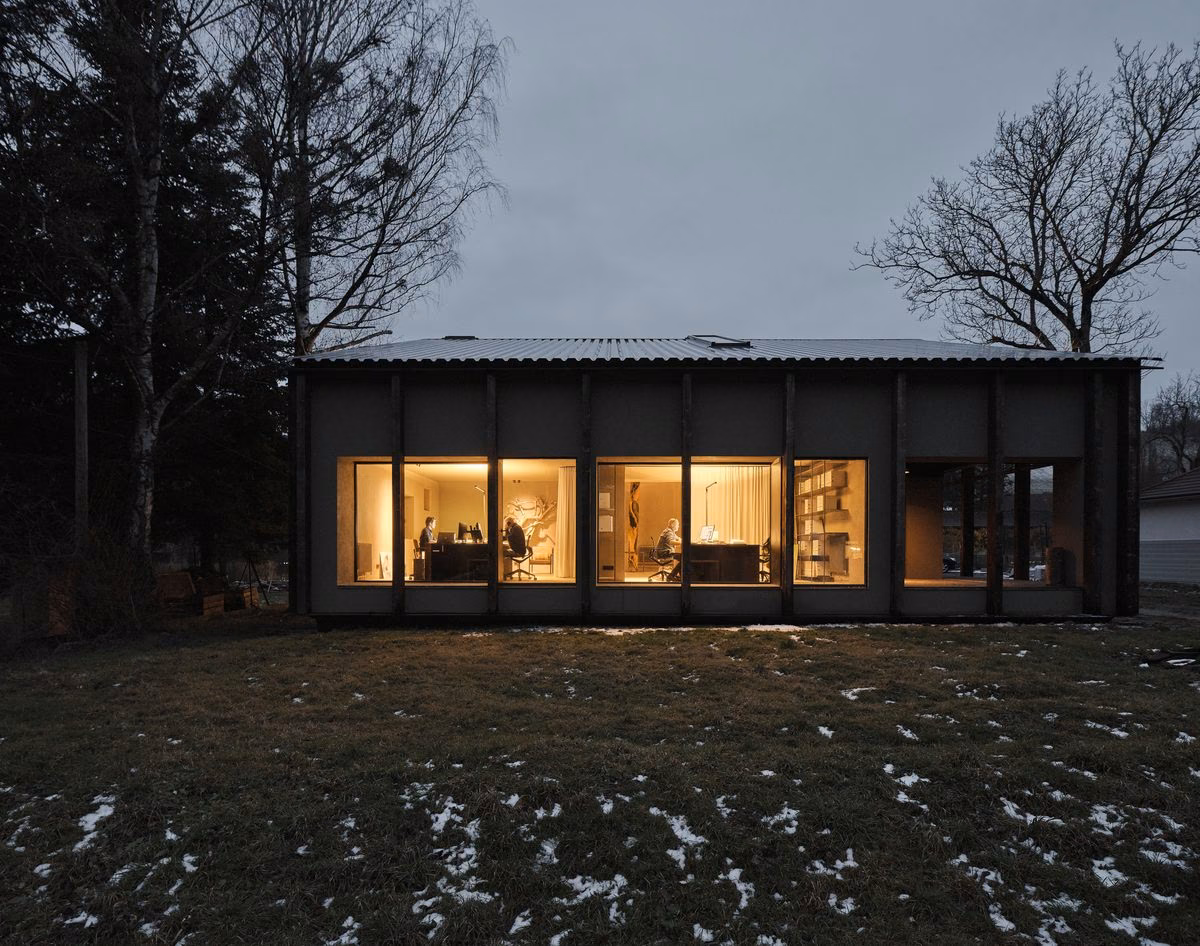 Das beleuchtete Atelierhaus am Abend, davor eine Wiese mit Schneeresten
